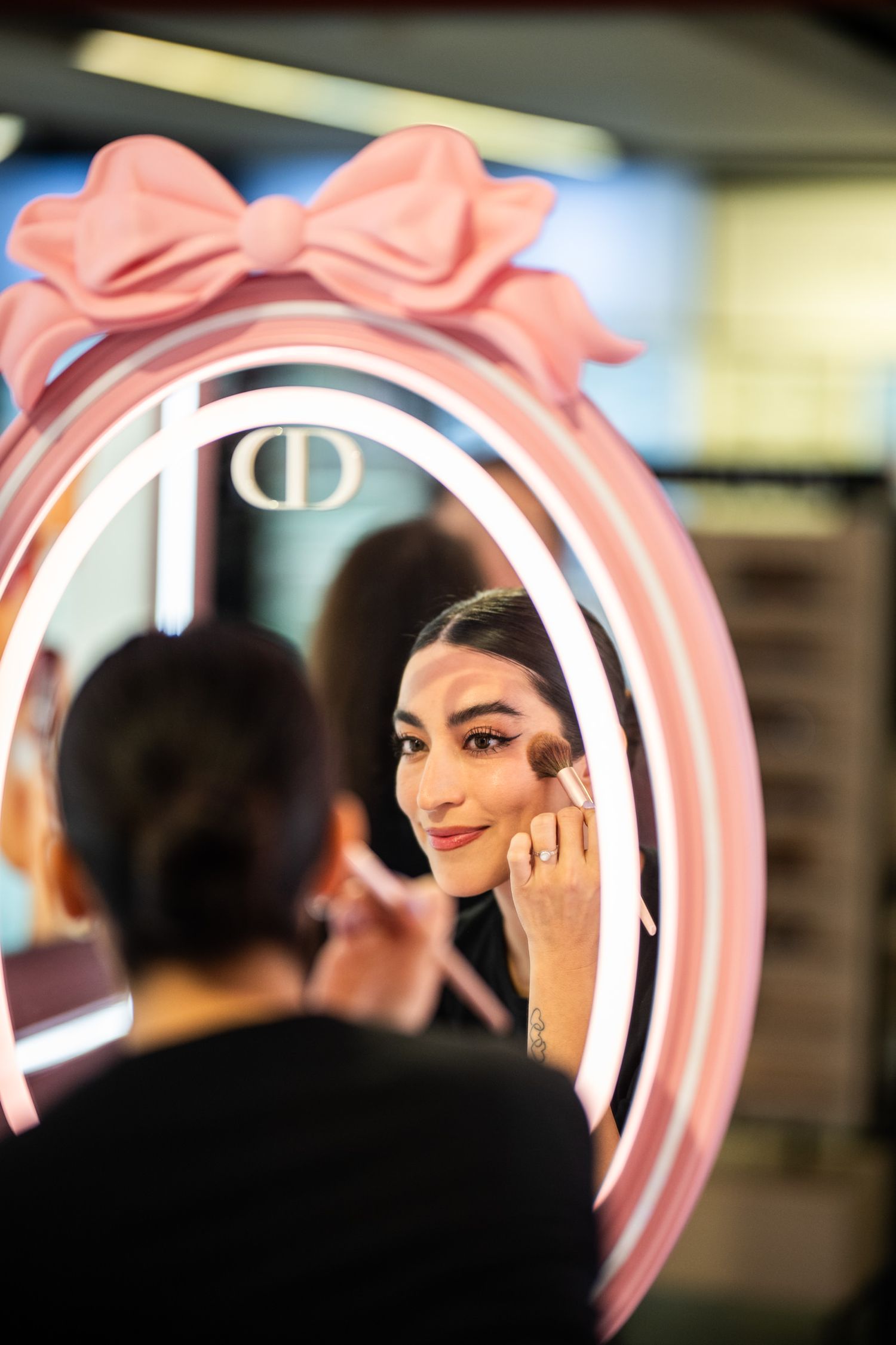 Woman applying makeup reflected in a pink oval Dior mirror with bow detail in a beauty store.