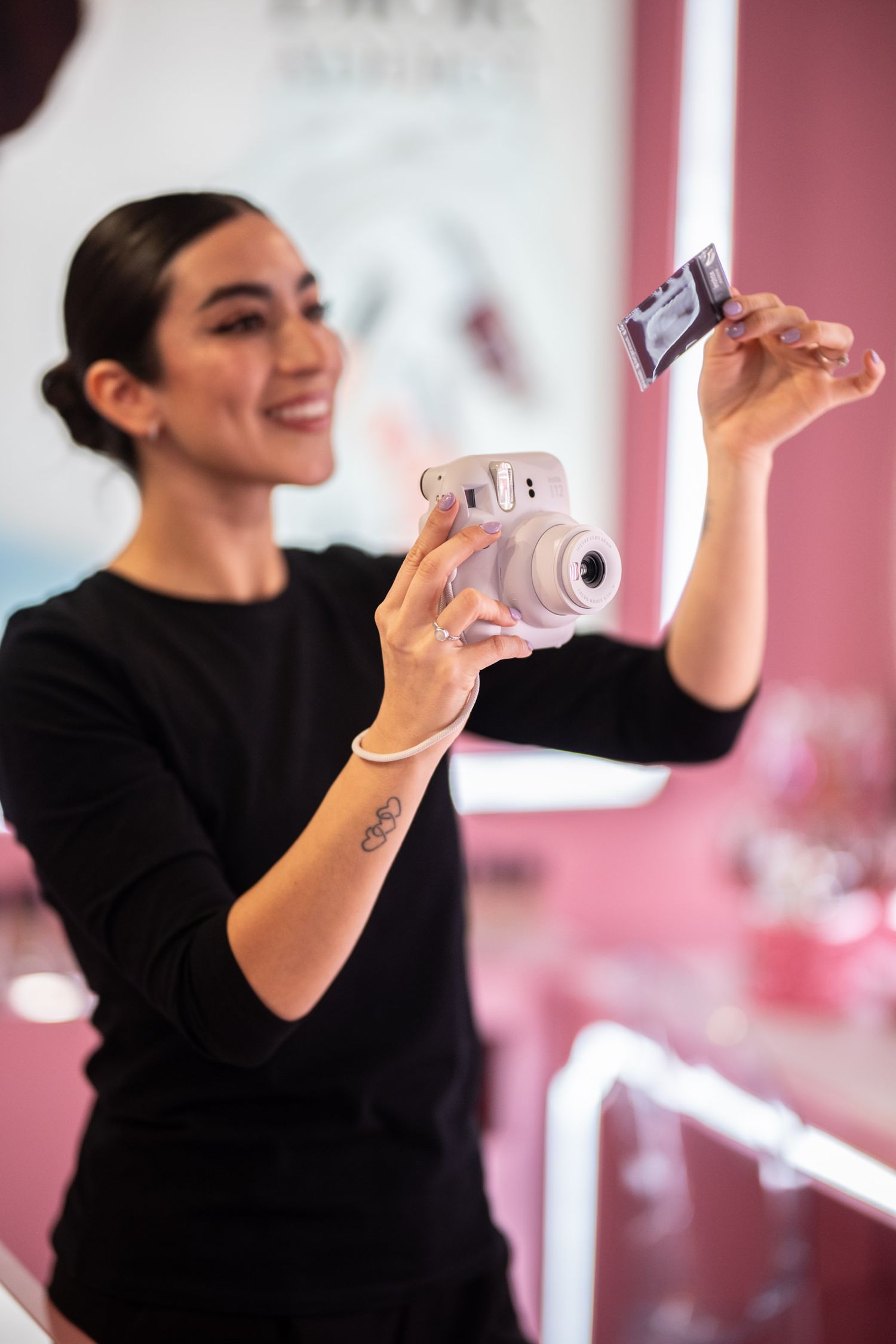Smiling woman in black top holding a Fujifilm Instax mini camera with a photo ejecting in a pink studio.