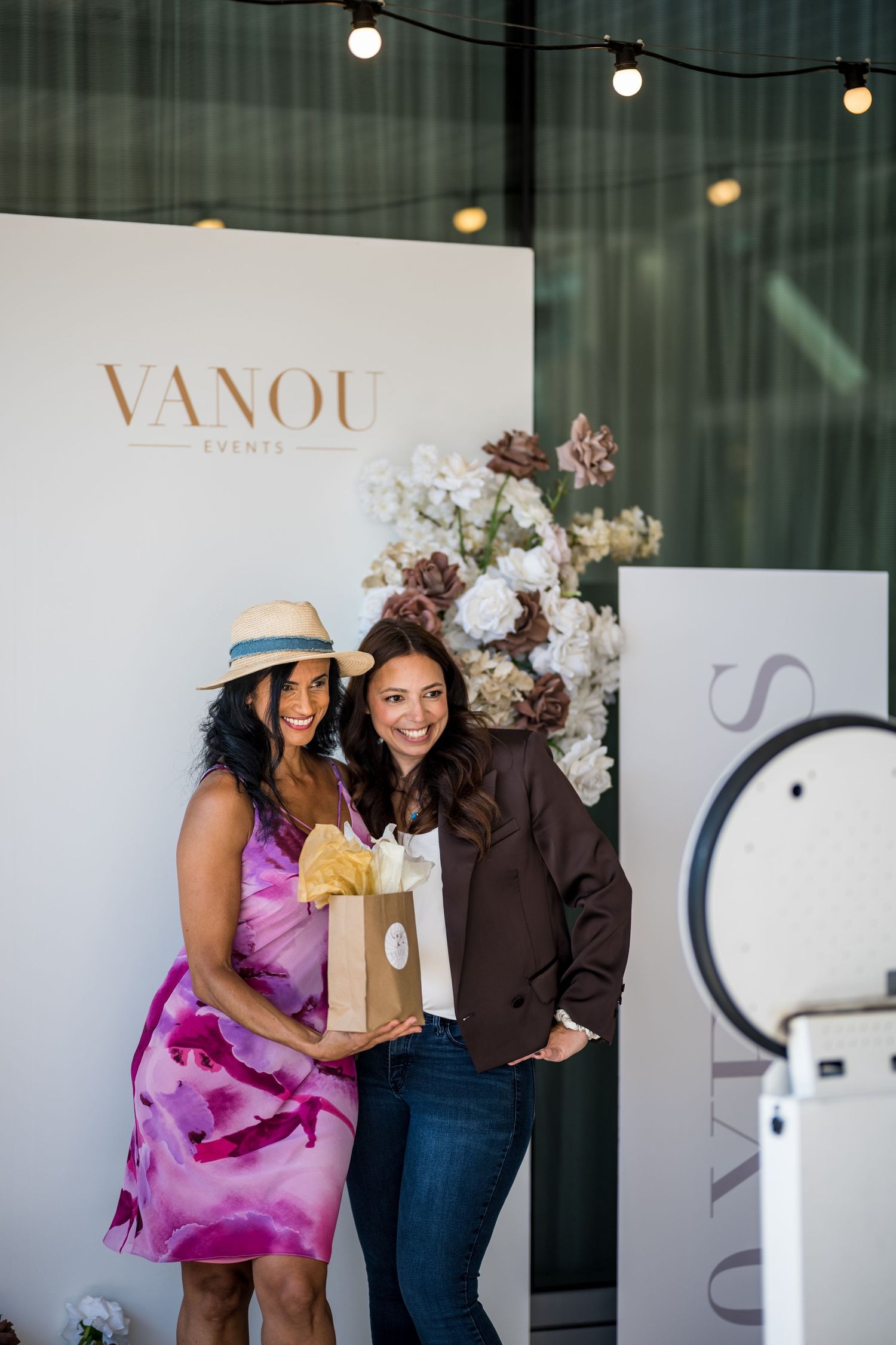 Two women smile and pose with gift bags in front of a Vanou Events branded backdrop with floral arrangements.