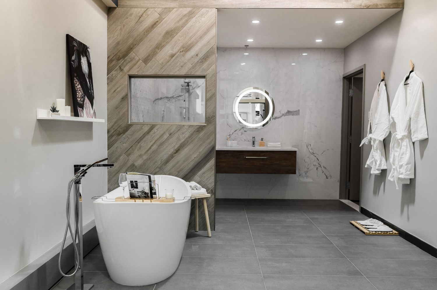 Modern luxury bathroom with freestanding white soaking tub, wood accent wall, round mirror, floating vanity, and white robes.