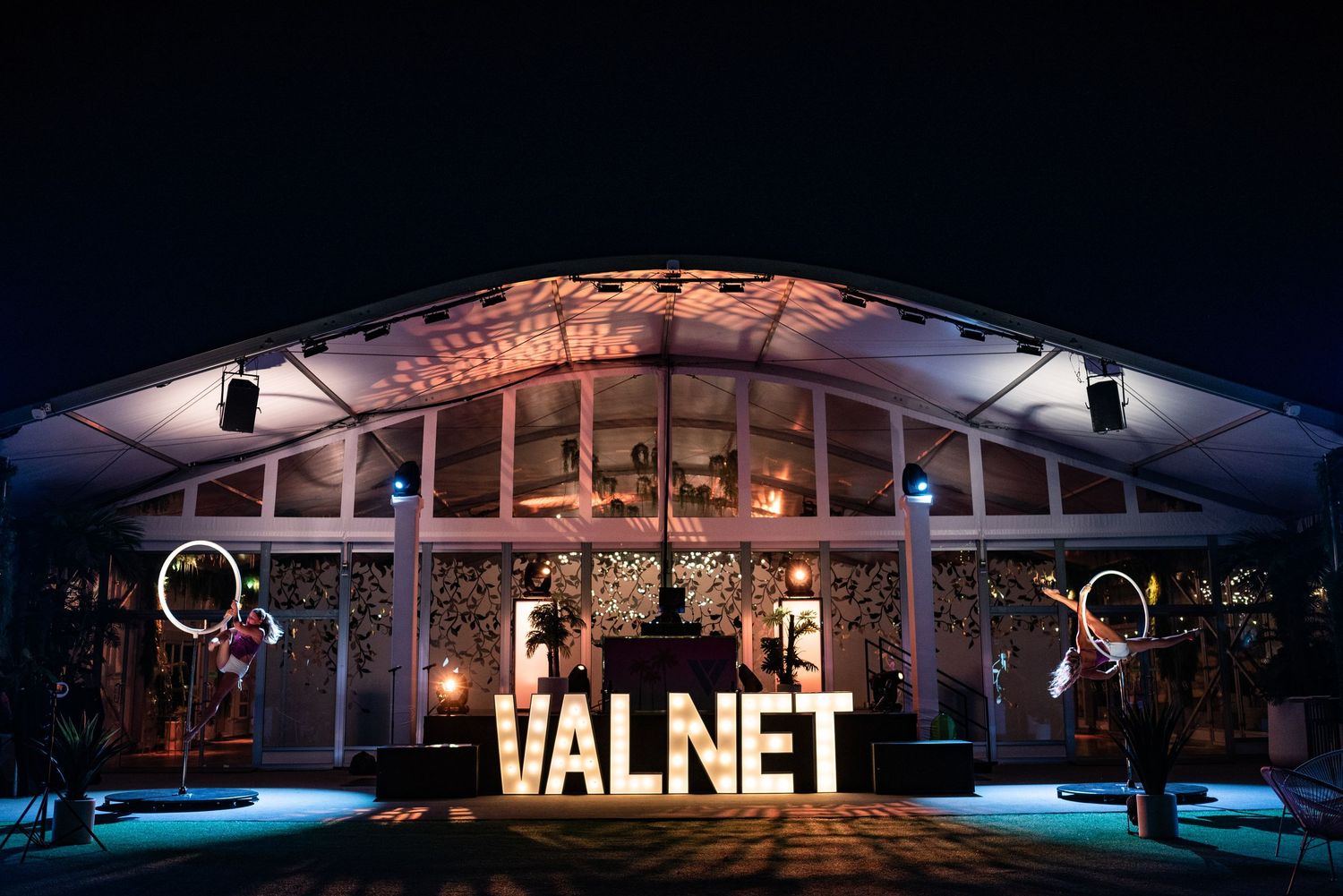 Large illuminated VALNET sign outside a marquee tent at a nighttime outdoor event with glowing hoop performers.