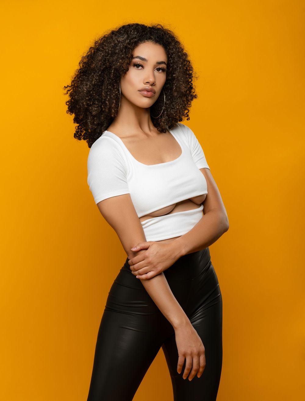 Creative studio portrait of a woman with curly hair wearing a white crop top against a bright yellow background.