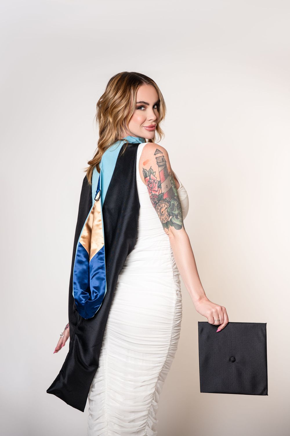 Fashion portrait of a blonde woman in a white fitted dress, holding a black shopping bag with colorful fabric draped over her shoulder.