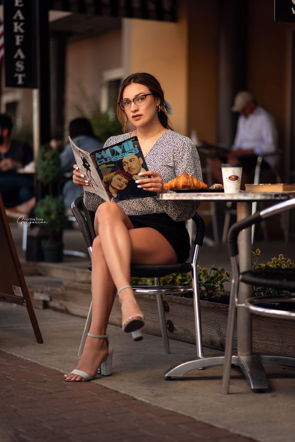 Lifestyle portrait of a stylish woman sitting at an outdoor café table reading a magazine.