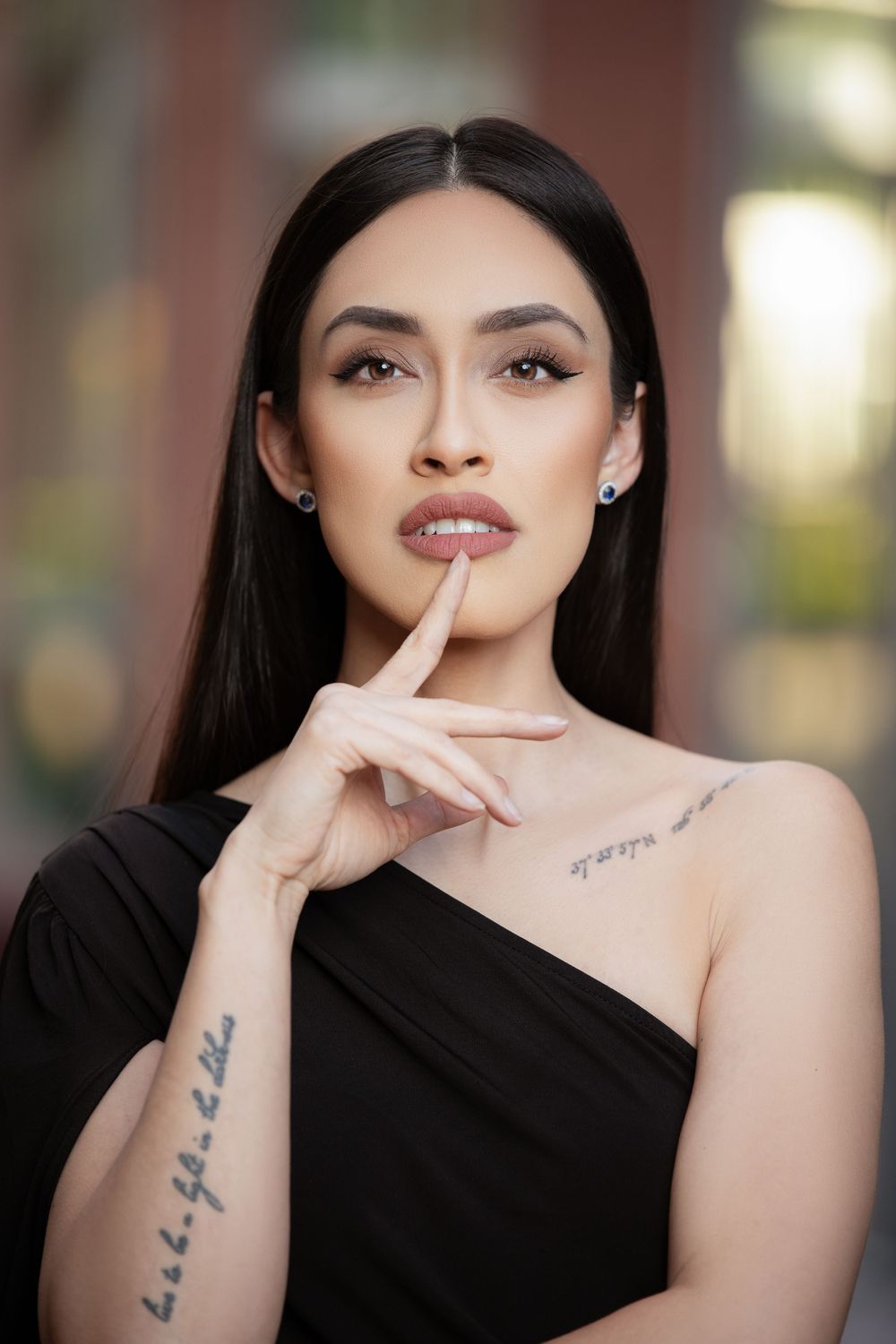 Close-up beauty portrait of a woman in a black one-shoulder top posing with her finger near her lips.