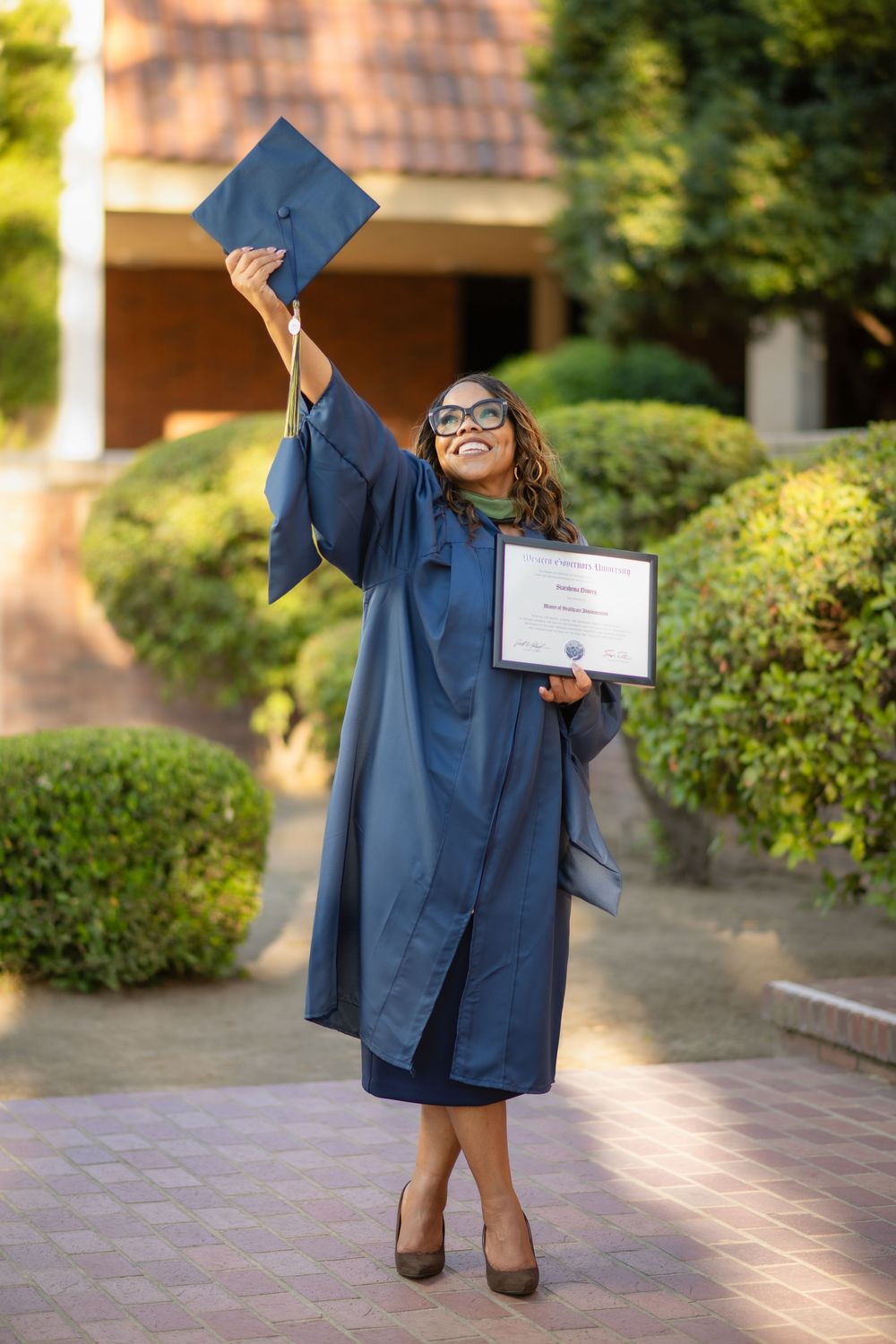 Smiling graduate in a blue cap and gown raising her graduation cap while holding a diploma outdoors.