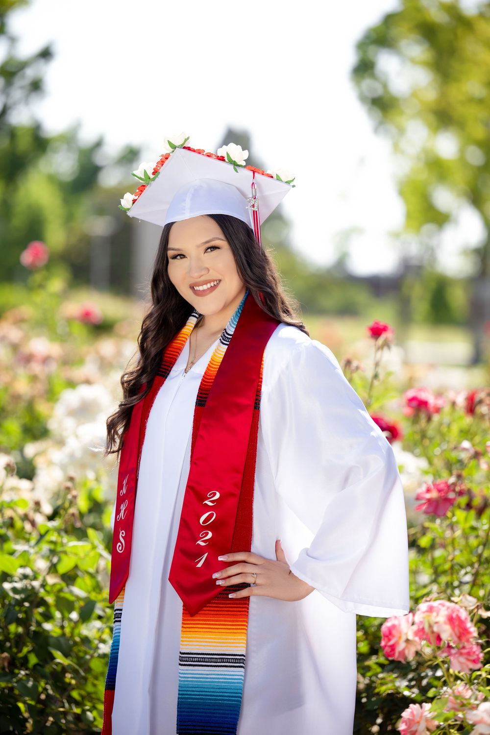 Graduate in a white cap and gown with a red stole posing among blooming flowers outdoors.