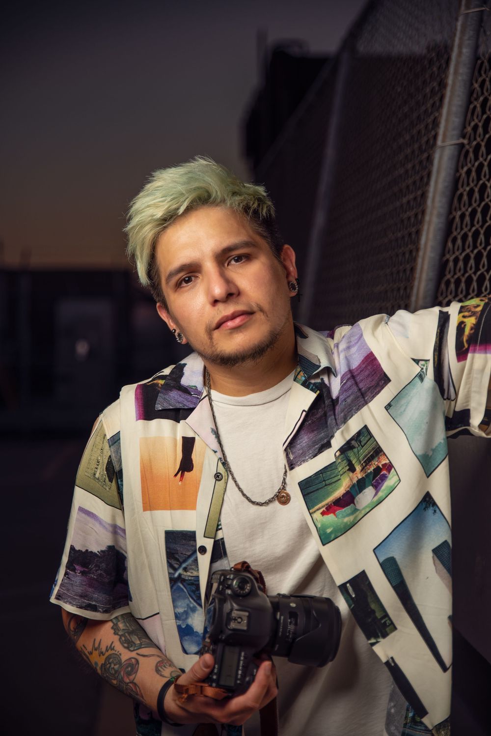 Creative portrait of a man with short blond hair holding a camera against a softly blurred urban background.