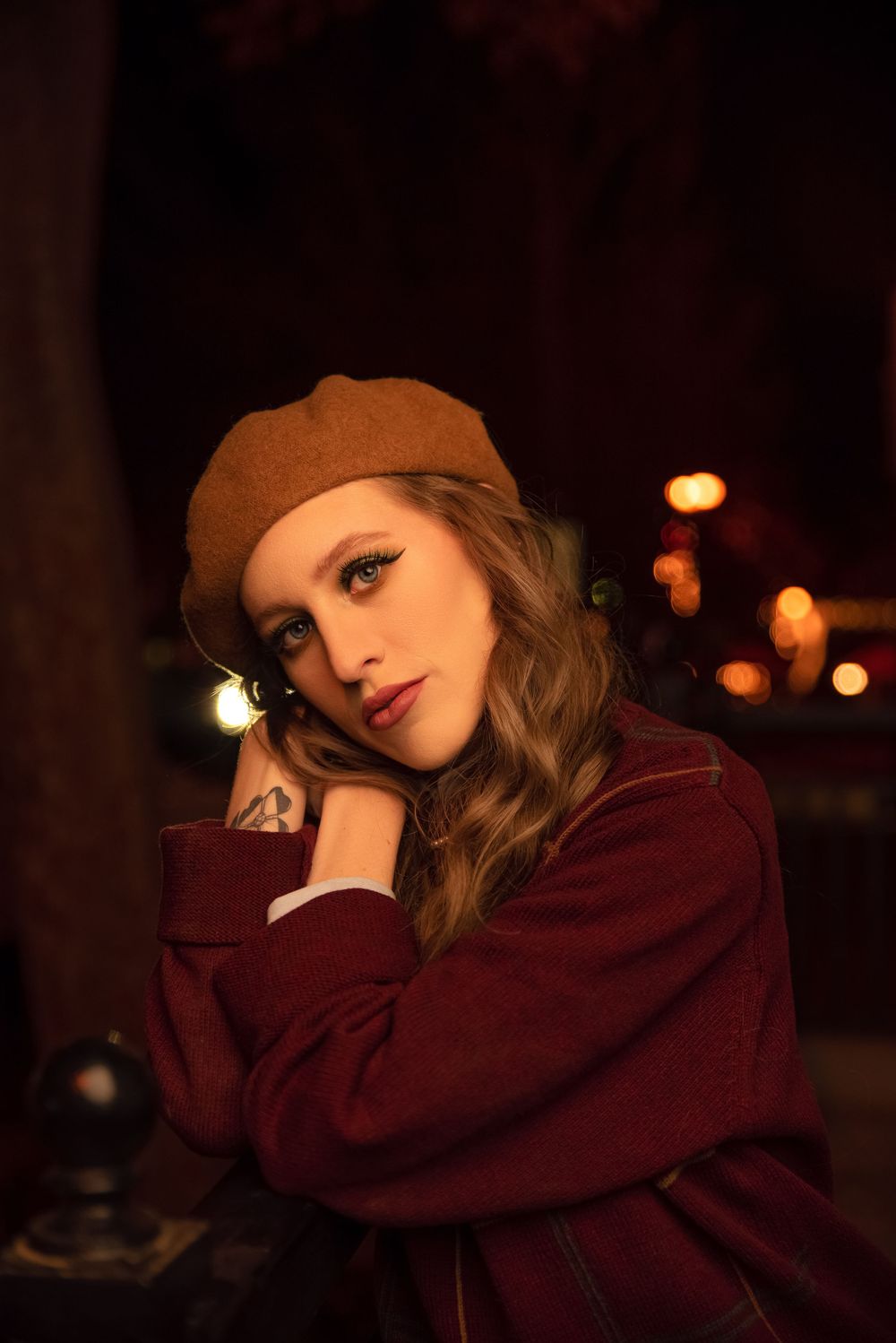 Moody nighttime portrait of a woman wearing a brown beret and burgundy sweater against warm city lights.