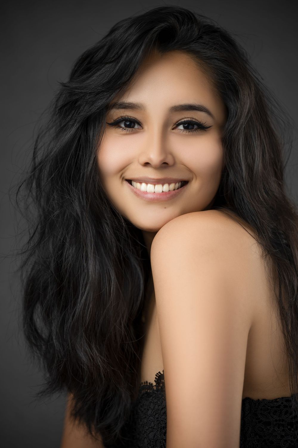 Smiling woman with long dark hair posing for a studio beauty portrait against a dark gray background.