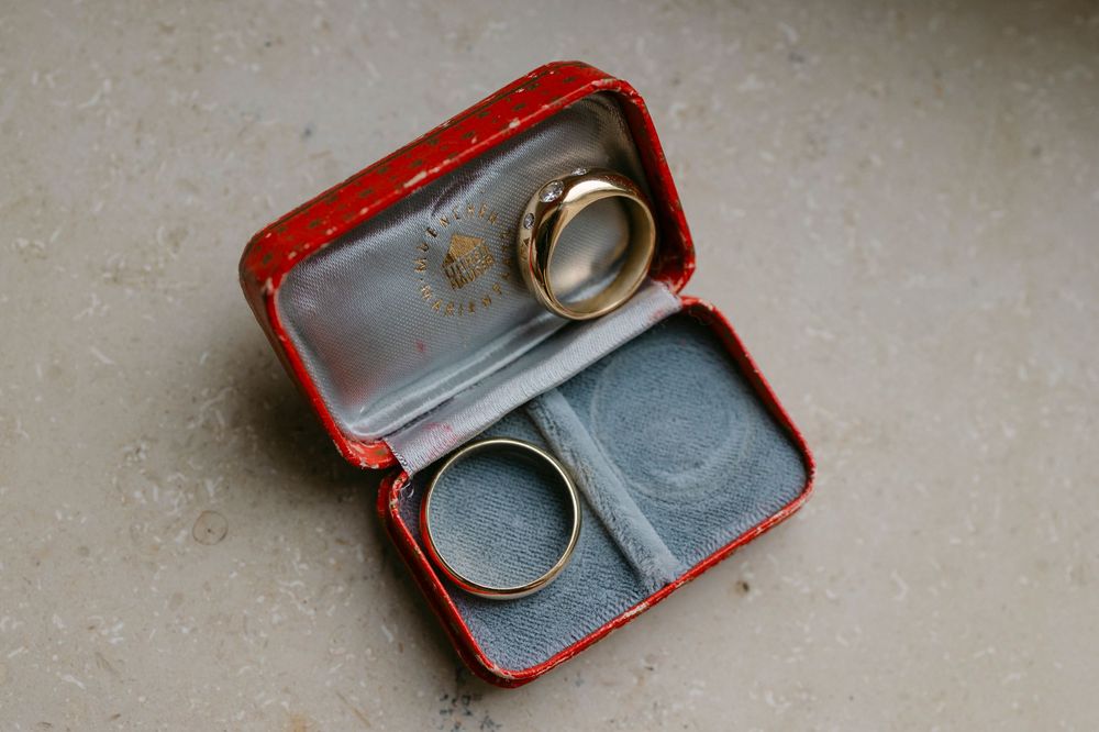 Two gold wedding bands resting in an open red jewelry box with a gray velvet interior, placed on a textured concrete surface.