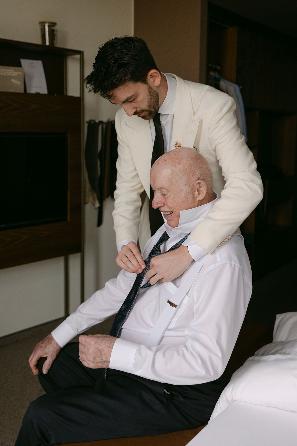 A groom in a cream jacket stands behind a seated older man in a white shirt, helping him adjust his tie in a hotel room.