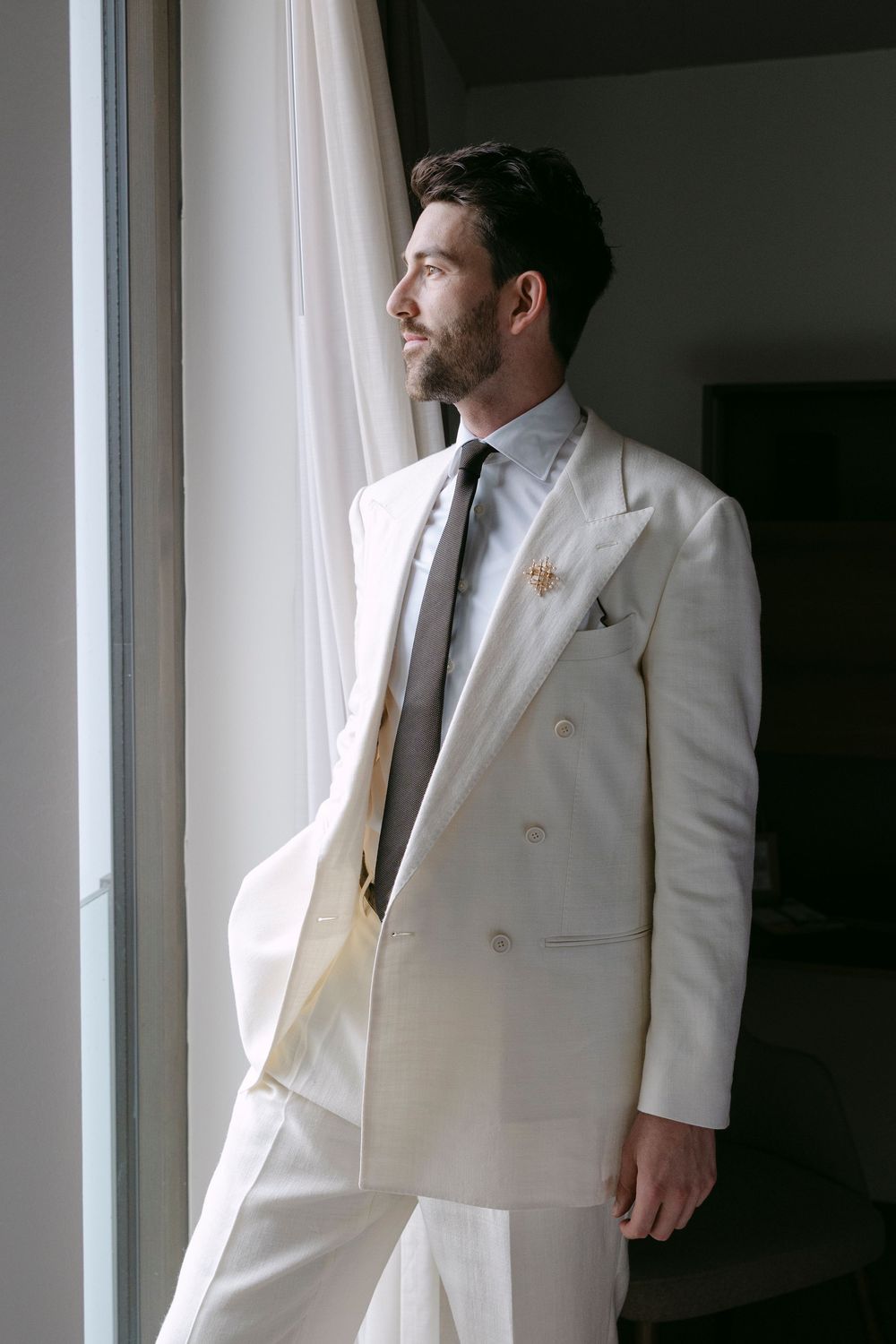 Bearded groom in a cream double-breasted suit and gray tie stands beside a sheer-curtained window, gazing outside in soft natural light.