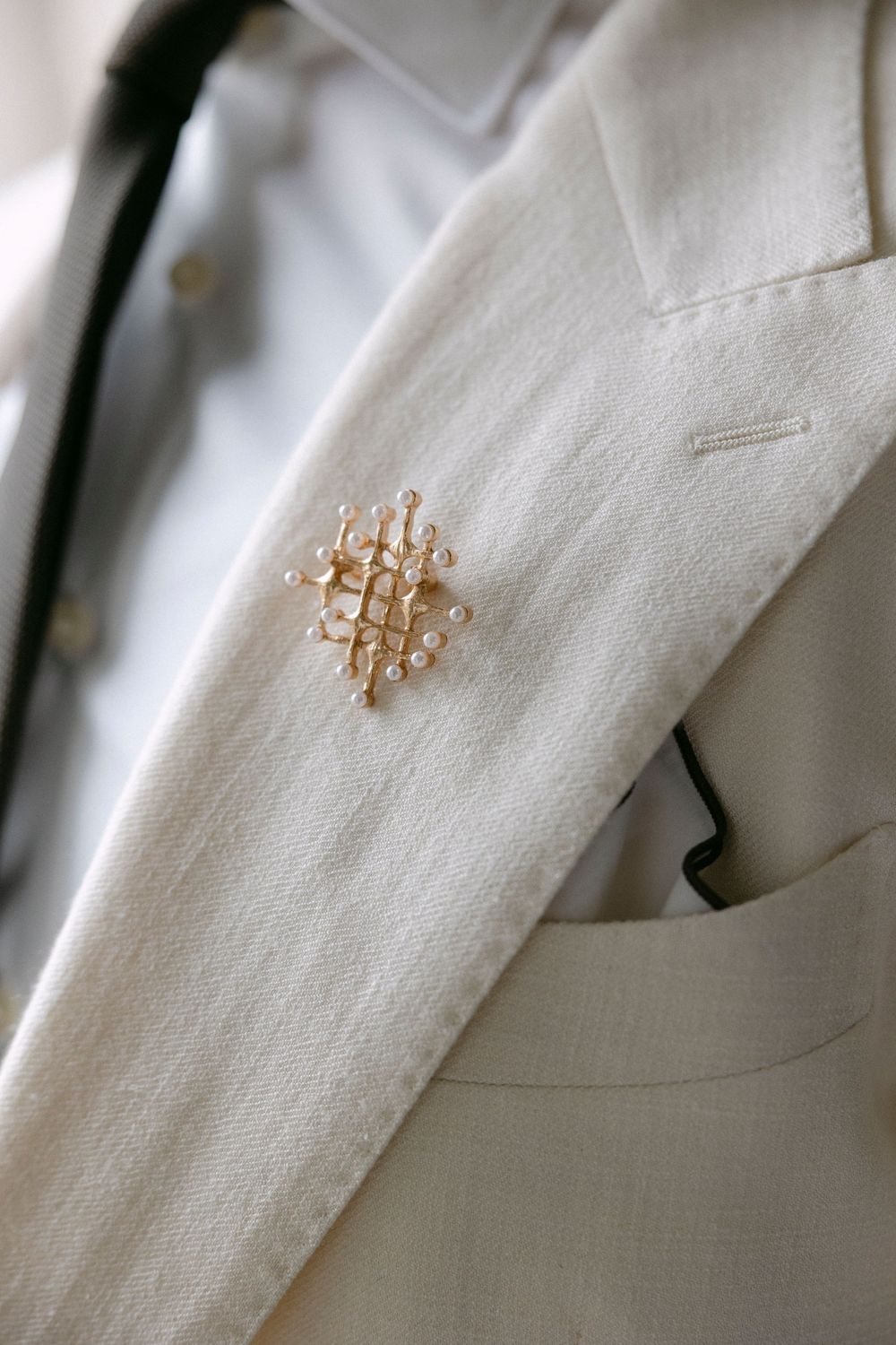 Gold pearl-studded lattice brooch pinned to the lapel of a cream linen suit jacket over a white shirt.