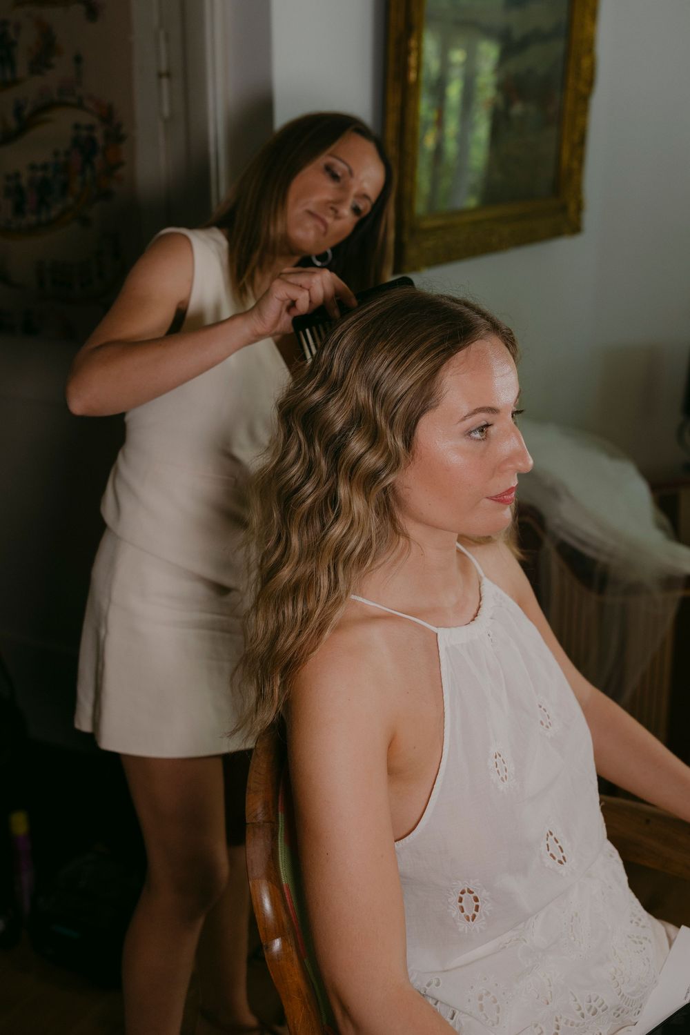 Hairstylist combs and styles the soft beach waves of a bride in a white embroidered halter dress while she gets ready for her wedding.