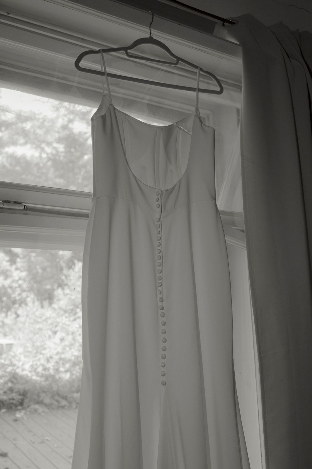 White wedding dress with thin straps and a row of fabric-covered buttons down the back hanging by a sunlit window.