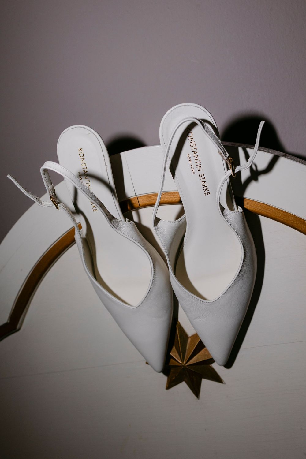 Pair of white pointed-toe slingback bridal heels by Konstantin Starke displayed on a round table with gold trim and star decor.