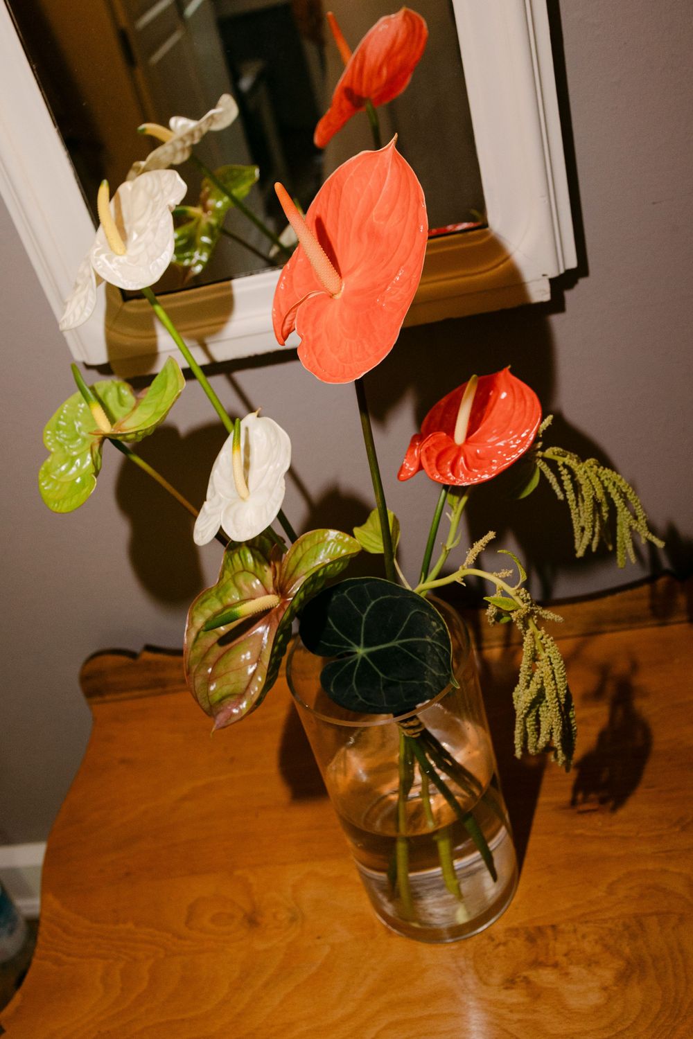 Red, white and green anthurium flowers arranged in a clear glass vase on a wooden table beside a mirror.
