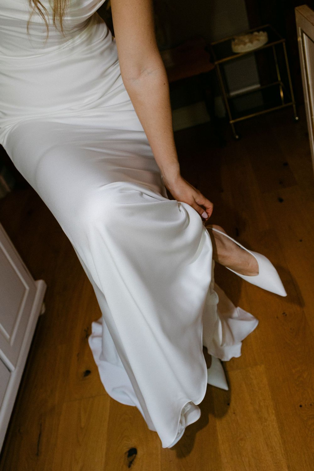 Bride in a flowing white satin wedding gown adjusts a pointed white bridal shoe while seated on a wooden floor indoors.