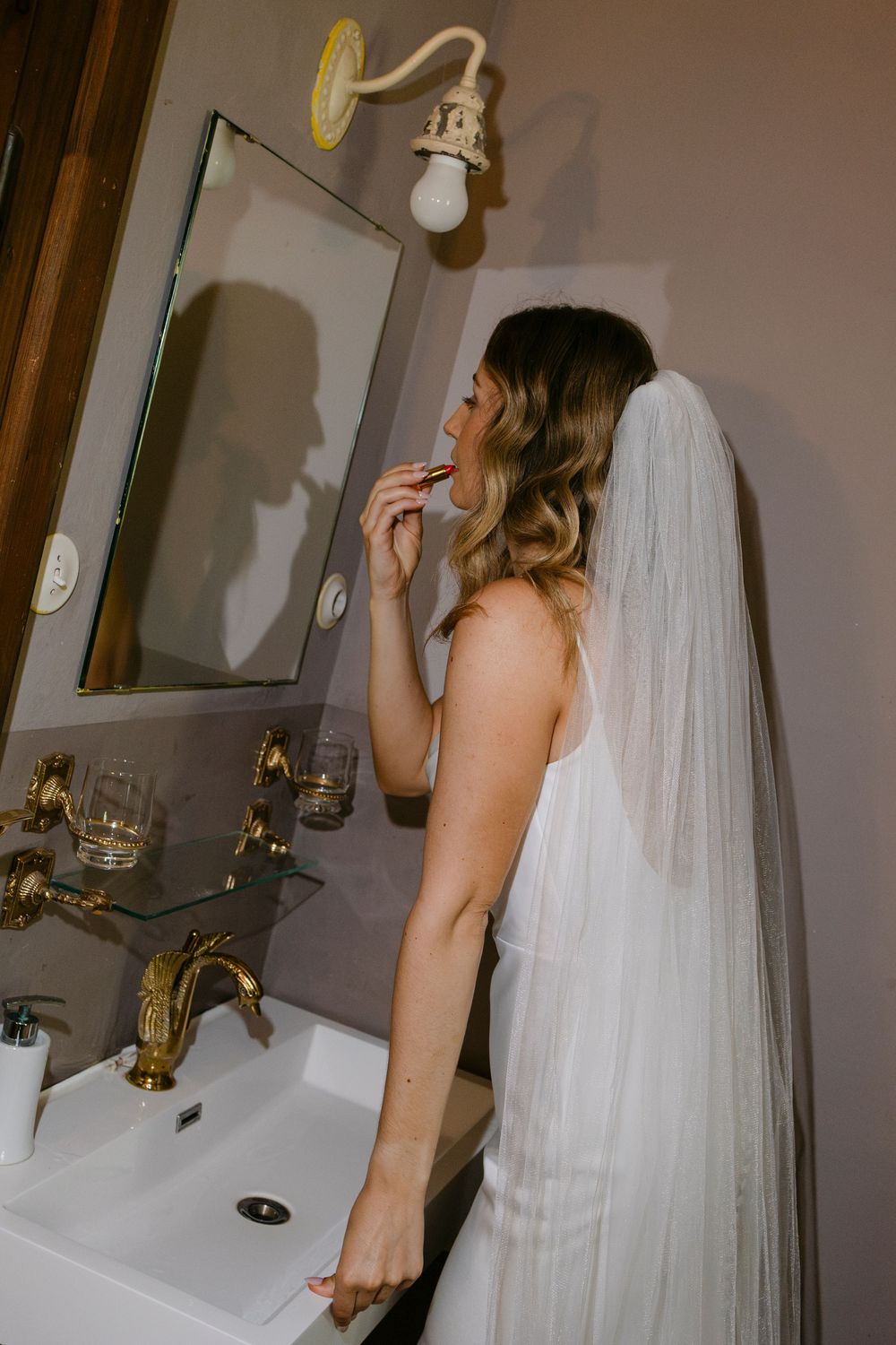 A bride in a white dress and long veil eats a snack while standing at a vintage bathroom sink with brass fixtures.