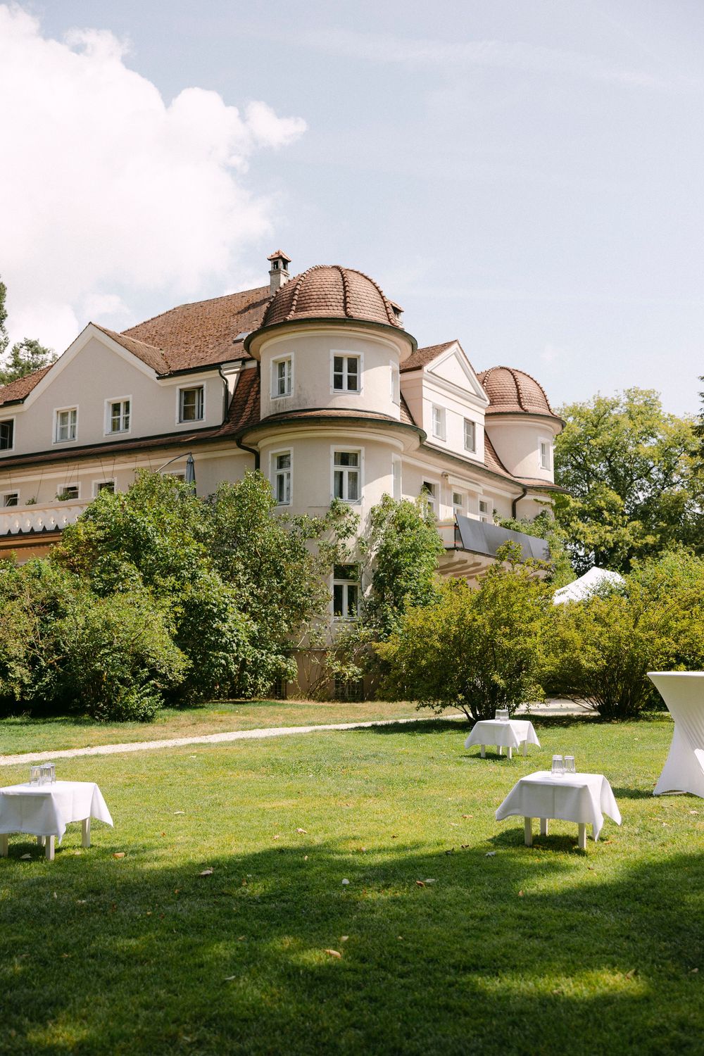 Elegant cream-colored manor with rounded turrets and a red tile roof overlooking a green lawn set with white cocktail tables.