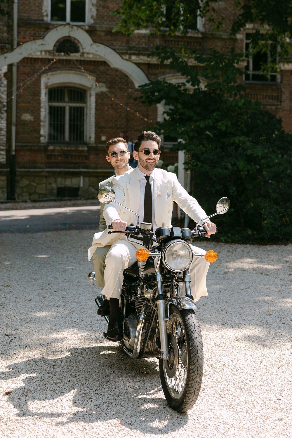 Two men in cream suits and sunglasses ride a vintage motorcycle along a gravel path in front of a historic red brick building.