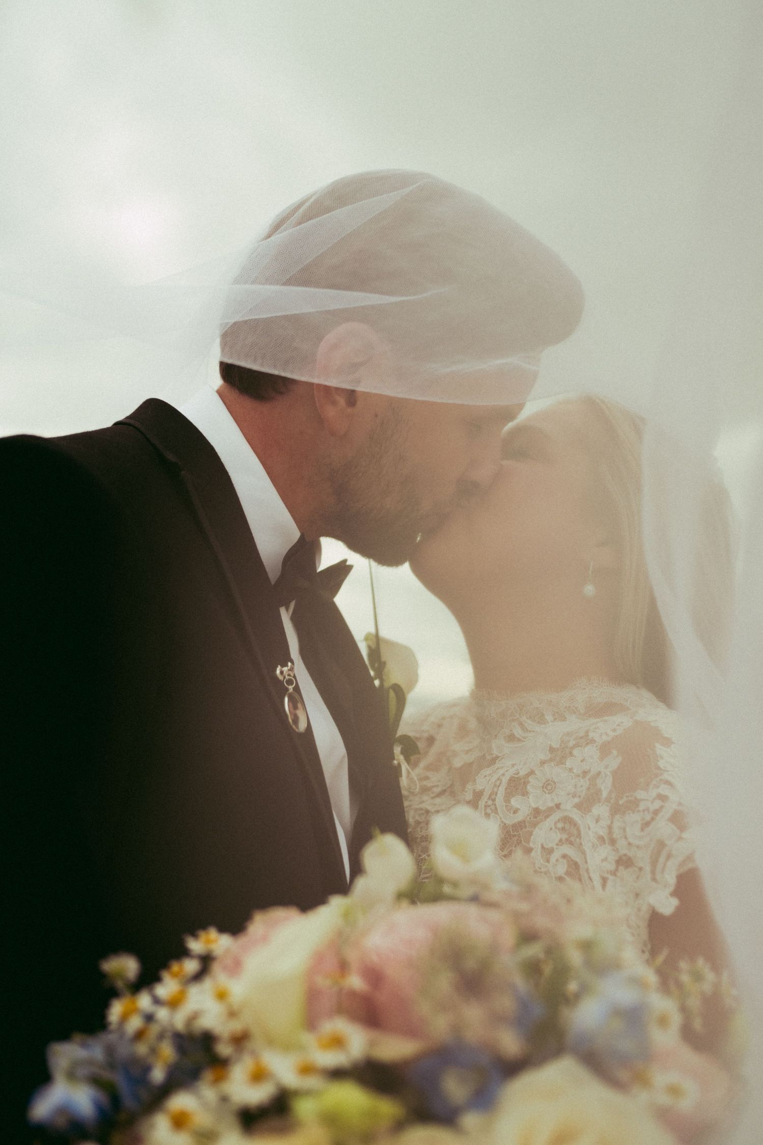 Bride and groom share a romantic kiss under a sheer veil, with a colorful pastel wedding bouquet in the foreground.