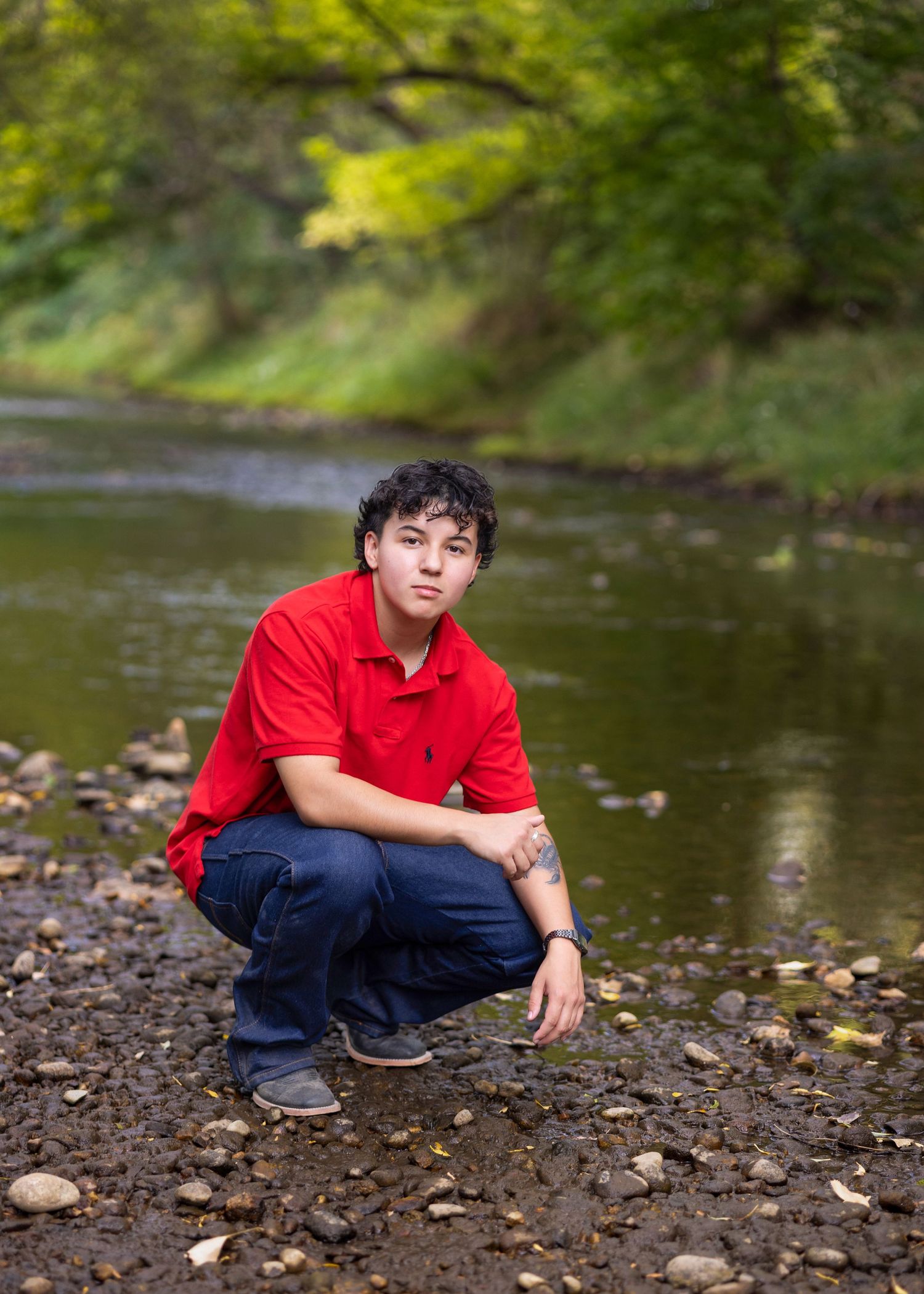 Young man in red polo shirt crouching by a rocky creek with lush green trees in the background.