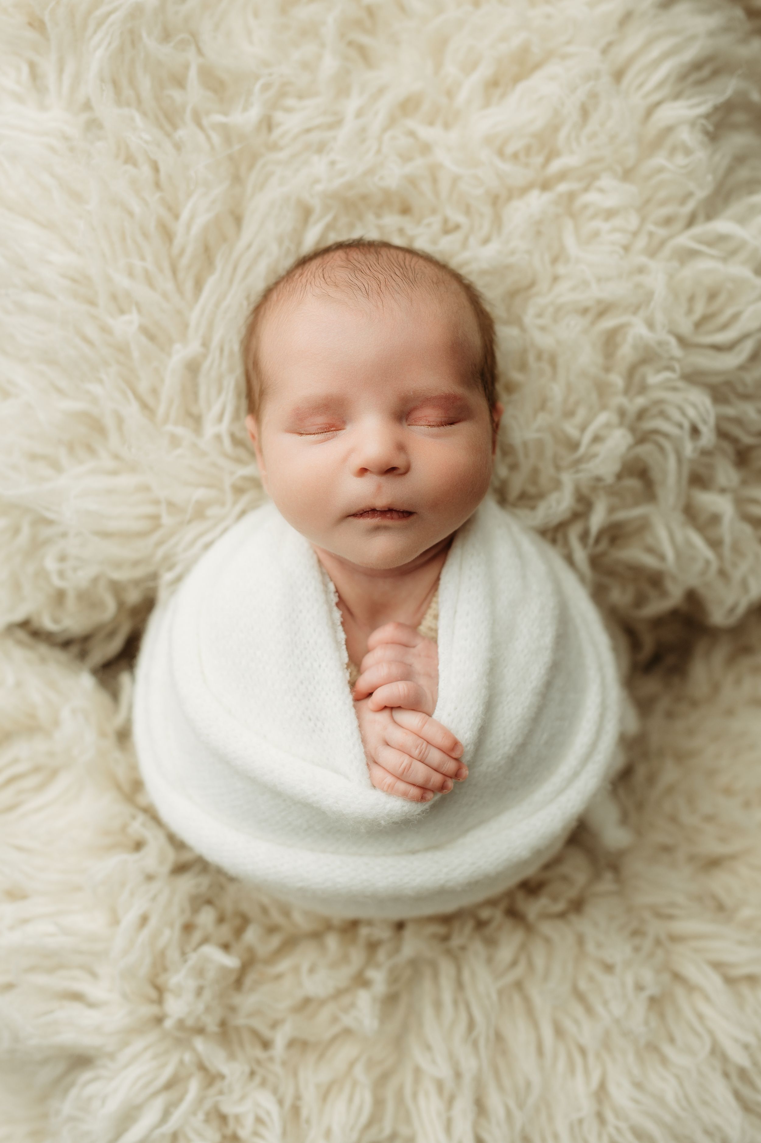 Peaceful newborn baby swaddled in white wrap sleeping on a fluffy cream fur blanket.