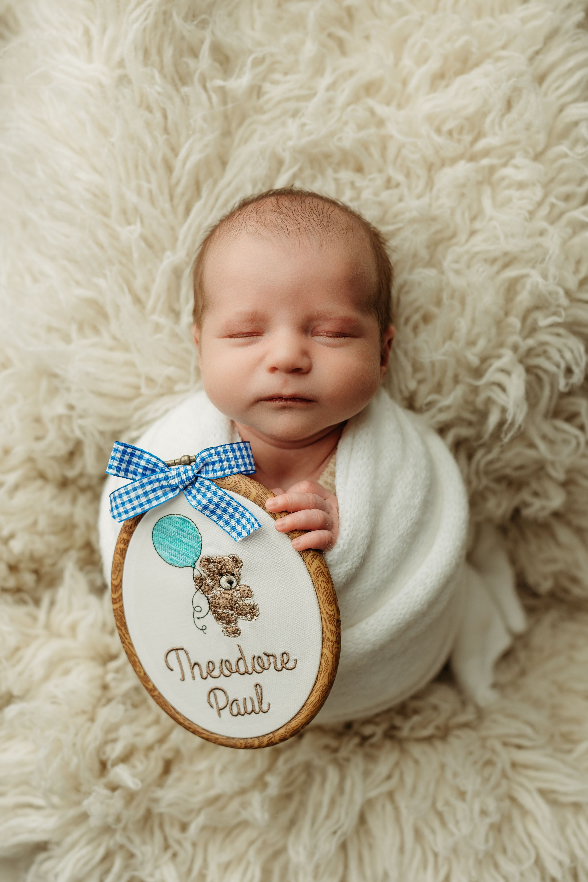 Sleeping newborn baby swaddled in white wrap holding a wooden name plaque reading Theodore Paul on a fluffy cream rug.
