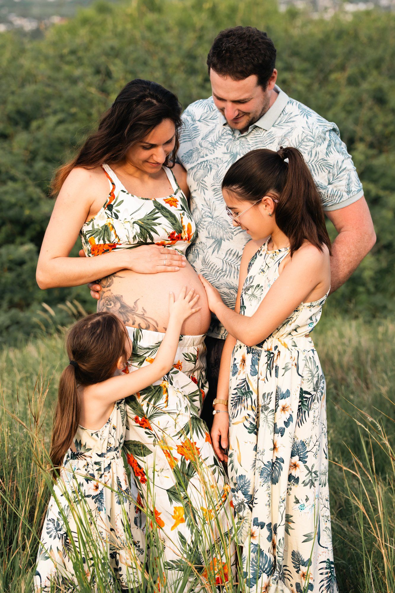 Pregnant woman with family touching baby bump outdoors in matching floral outfits.