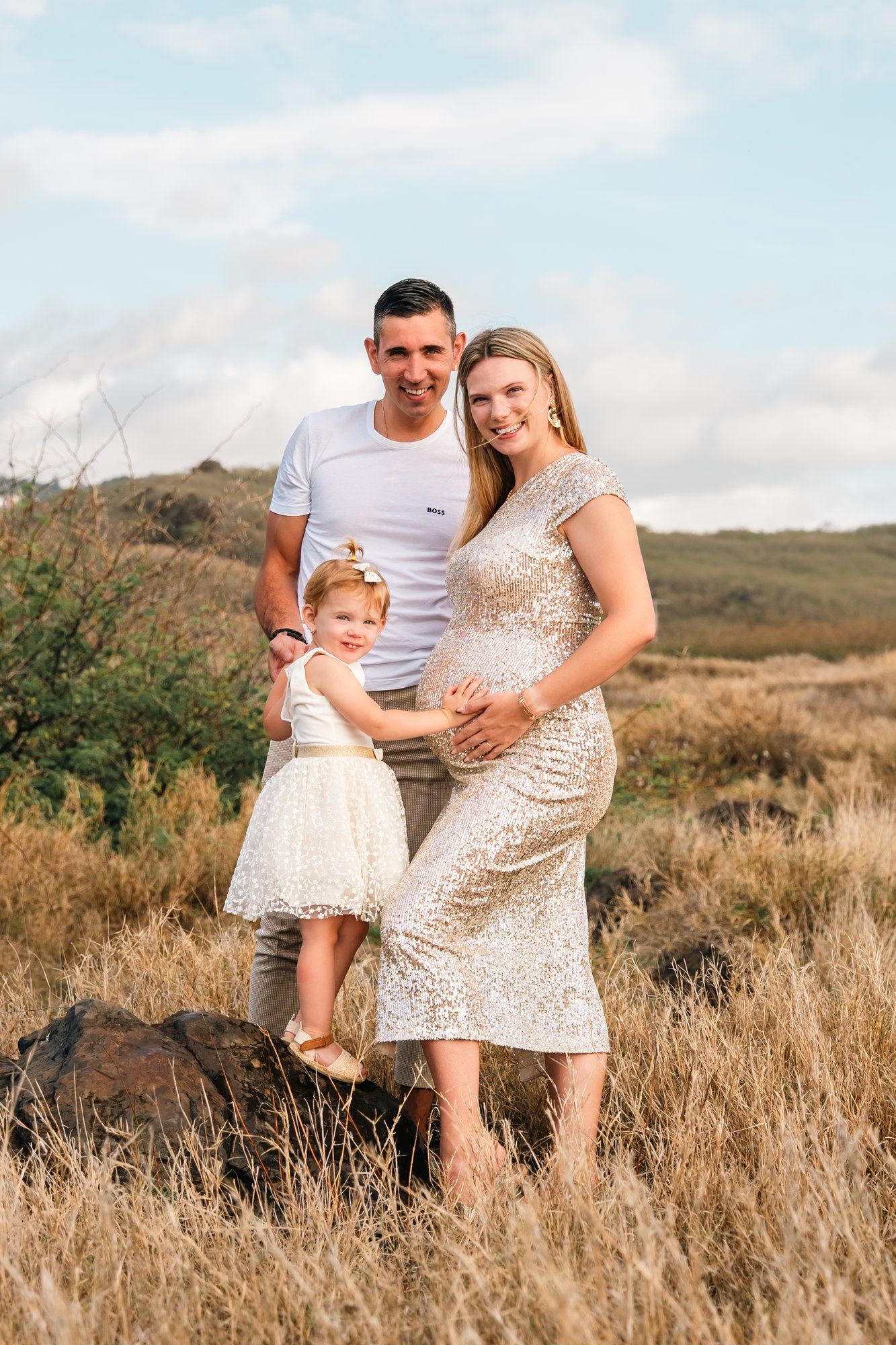 Pregnant woman in sequin dress with husband and toddler daughter posing in golden field for maternity photo.