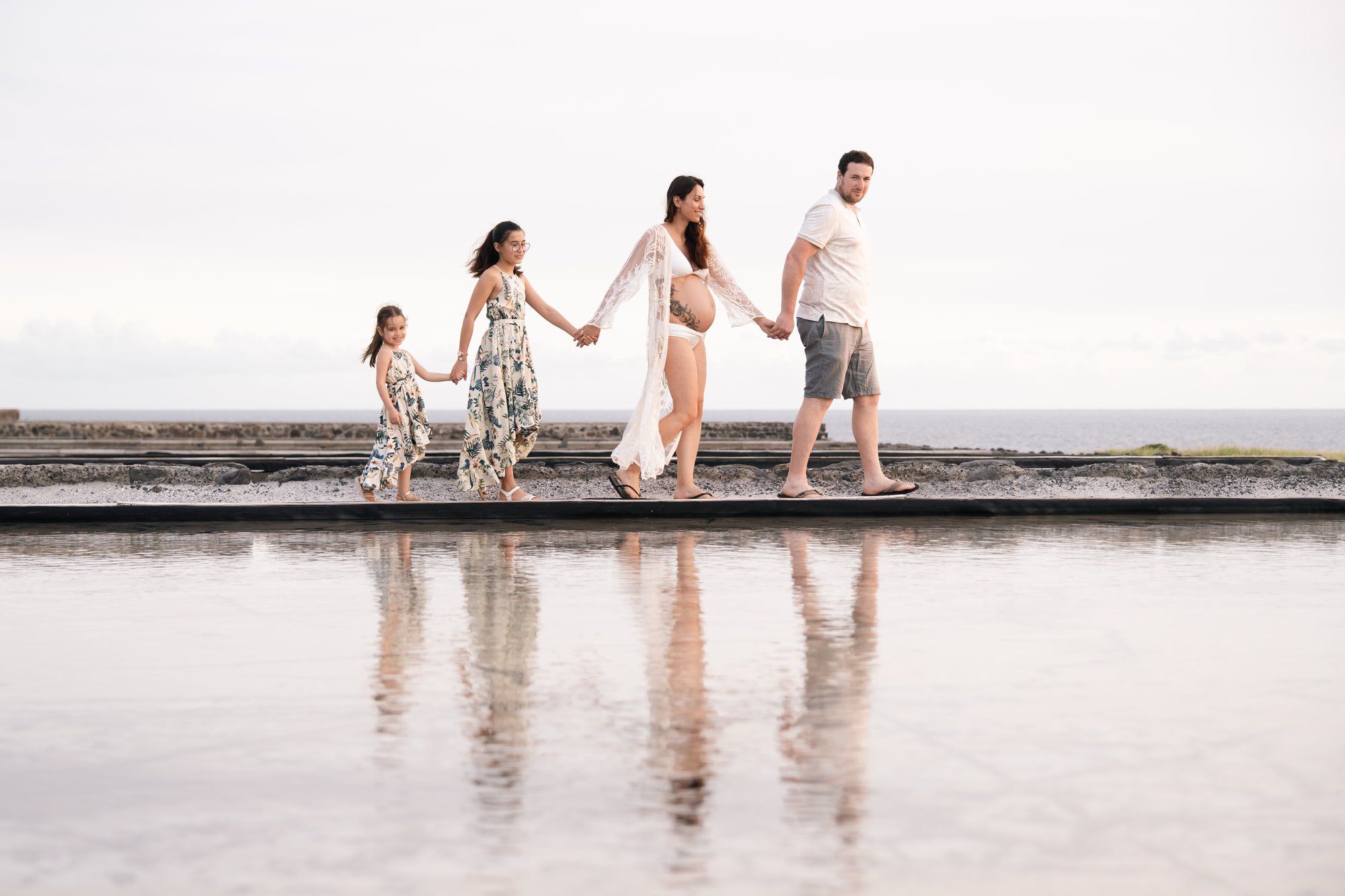 Family of four holding hands walking along reflective coastal boardwalk during maternity photoshoot.