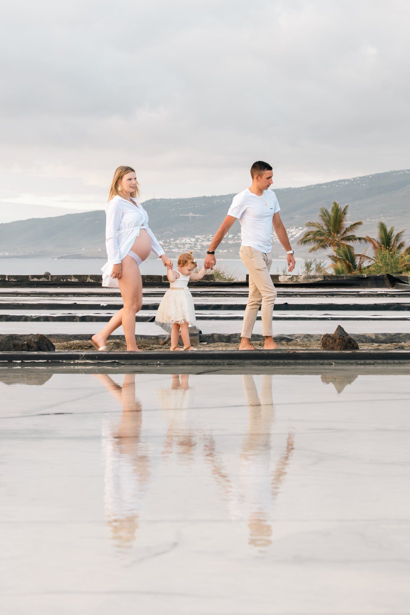 Pregnant woman and man walk with toddler on reflective surface near ocean.