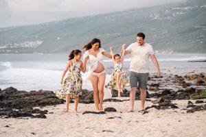 Pregnant woman and family joyfully running on sandy beach with ocean waves and green hills in background.