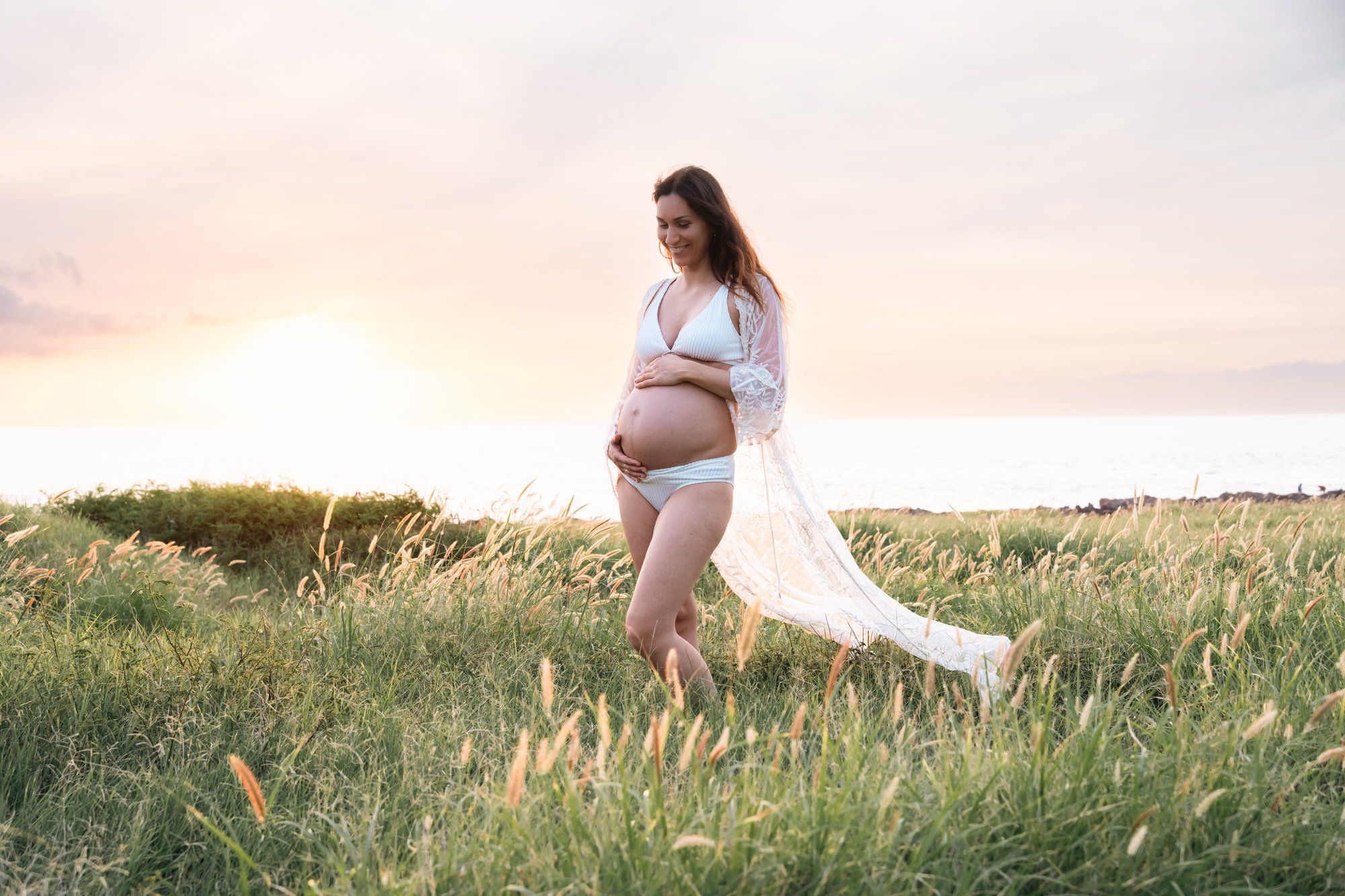 Pregnant woman in white lingerie and flowing cape stands in golden field at sunset, cradling her baby bump.