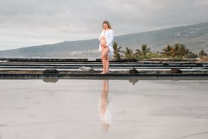 Pregnant woman in white dress stands on reflective surface near ocean with palm trees and mountains backdrop.