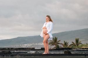 Pregnant woman in white shirt poses on a pier with tropical mountains and cloudy sky in Hawaii.