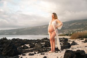 Pregnant woman in white lace robe poses on rocky Hawaiian beach with ocean and mountains backdrop.
