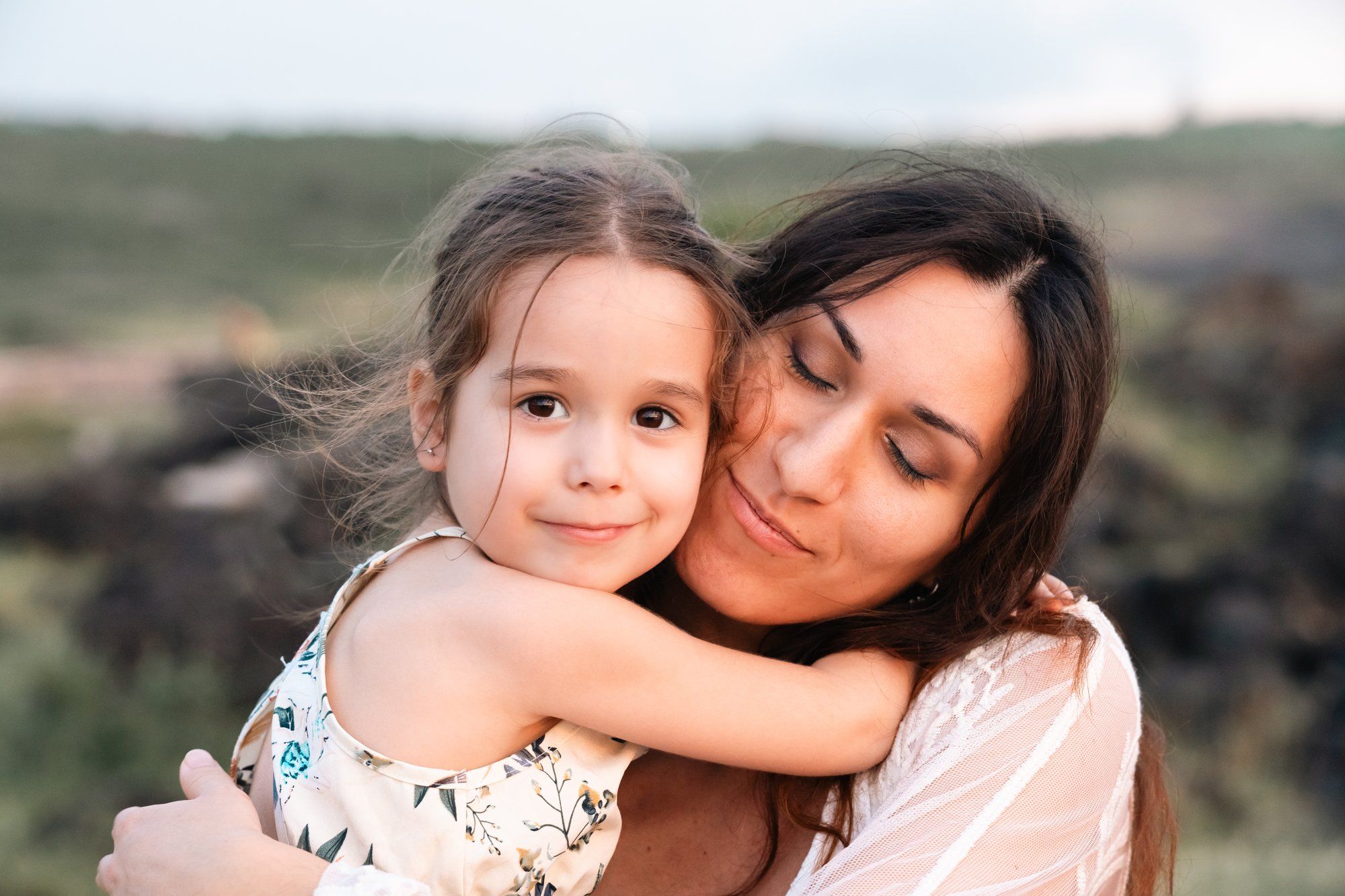 A mother tenderly embraces her young daughter outdoors, both sharing a warm and loving moment in a green field.