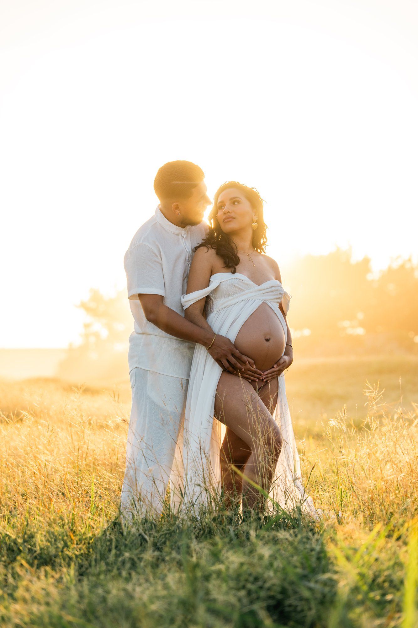 Couple in white embracing in golden sunset field, pregnant woman cradling baby bump, romantic maternity photo outdoors.