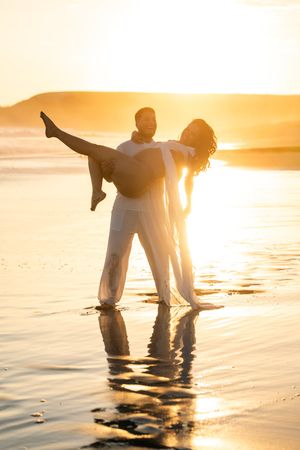 Man carrying woman in his arms on a beach at golden sunset, both dressed in white, with water reflections.