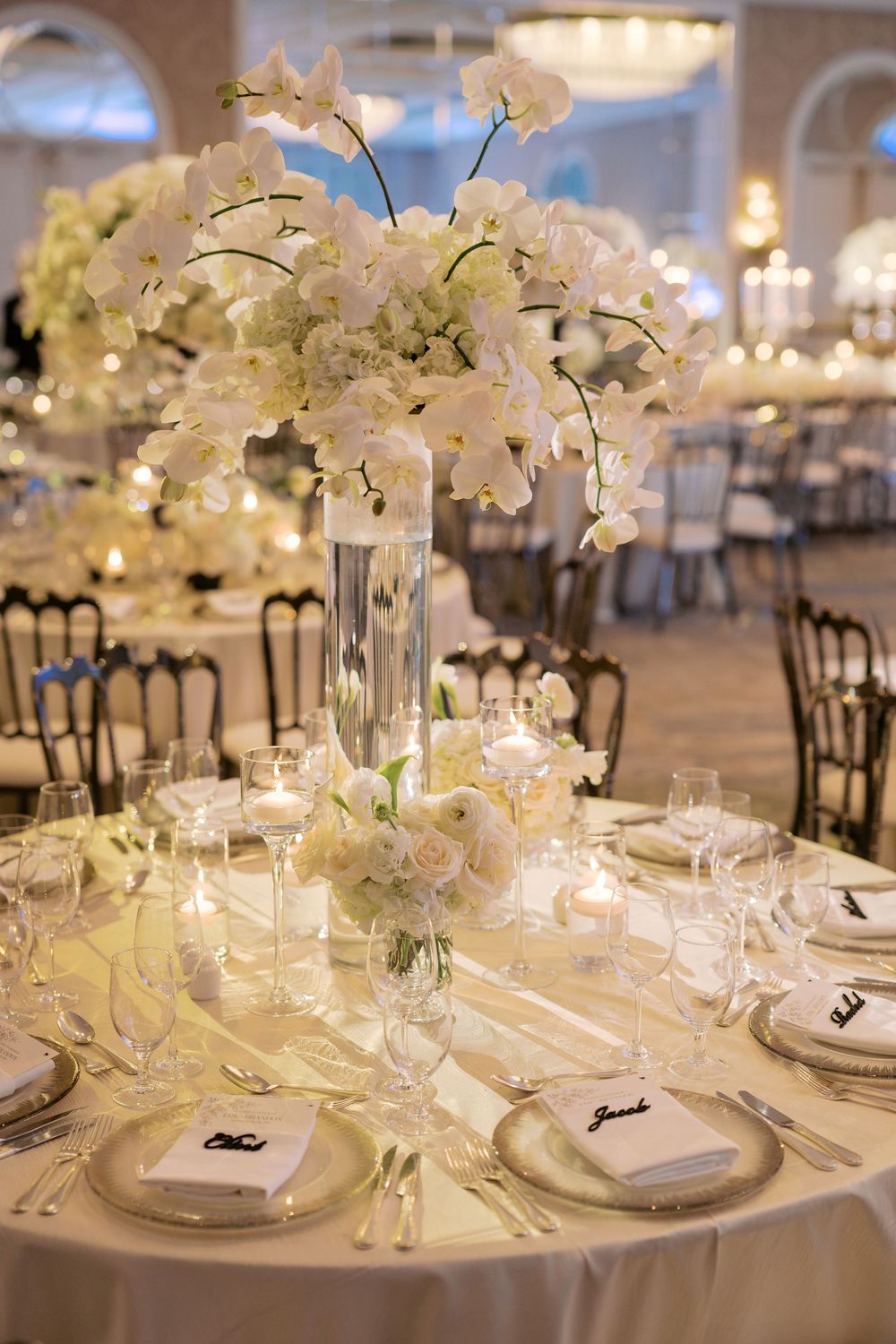 Elegant wedding reception table with tall white orchid centerpiece, crystal glasses, and ivory linens in a grand ballroom.