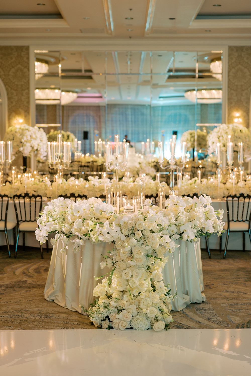 Elegant wedding reception with lush white floral centerpieces, candles, and ivory draping in a luxurious ballroom.