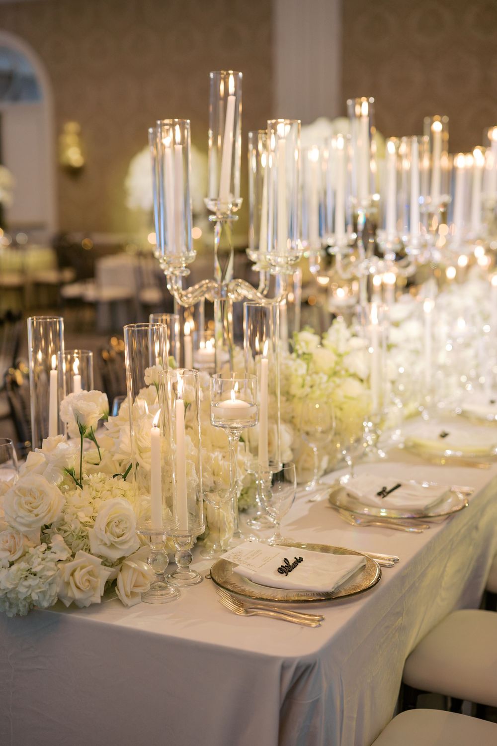 Elegant wedding reception table with white roses, hydrangeas, tall crystal candelabras, and glowing candles on a white linen.