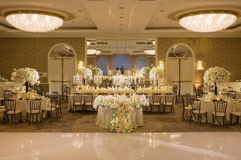 Elegant wedding reception ballroom with gold chairs, white floral centerpieces, crystal chandeliers, and a grand sweetheart table.