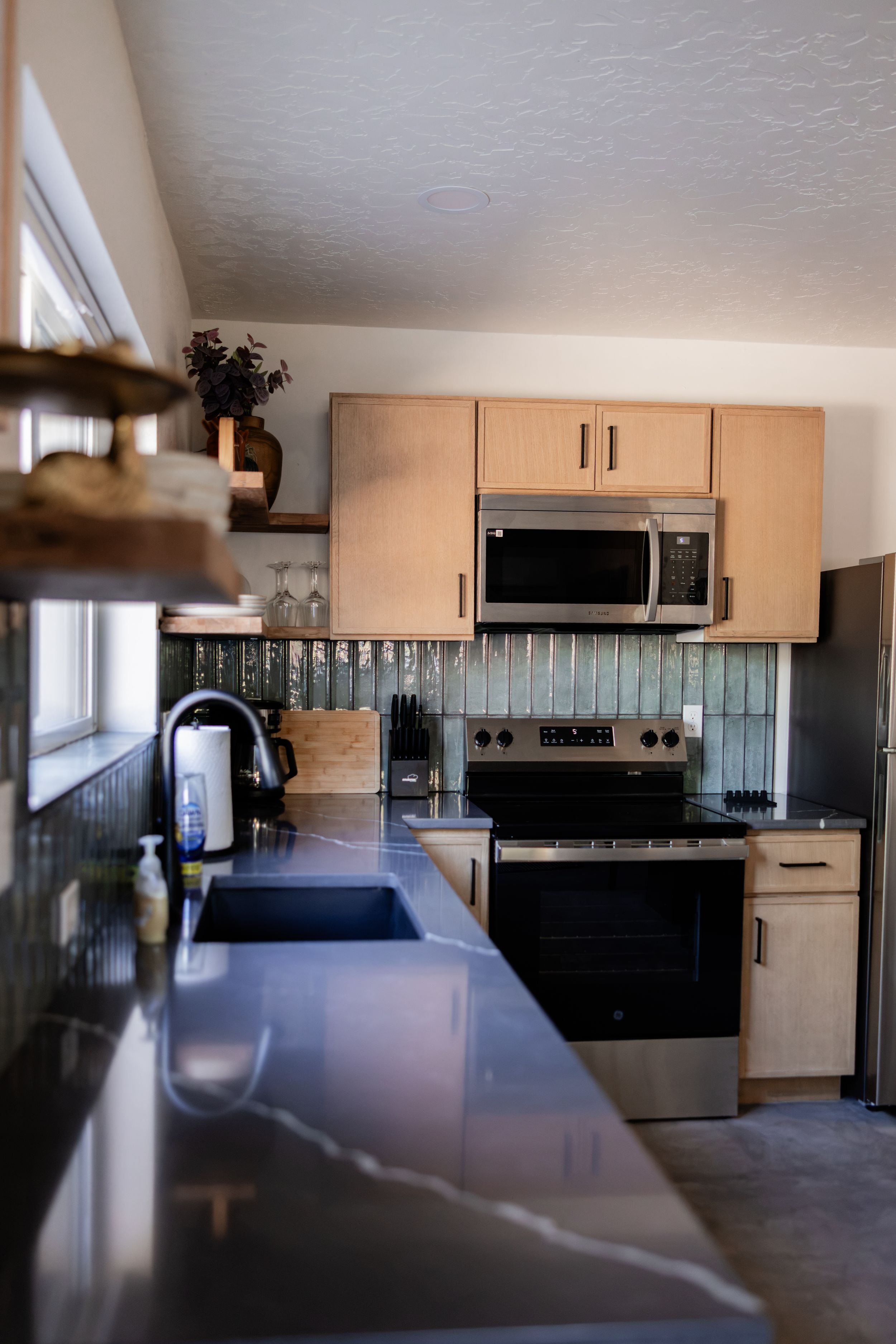 Modern kitchen with stainless steel appliances, light wood cabinets, dark countertops, and metallic tile backsplash.