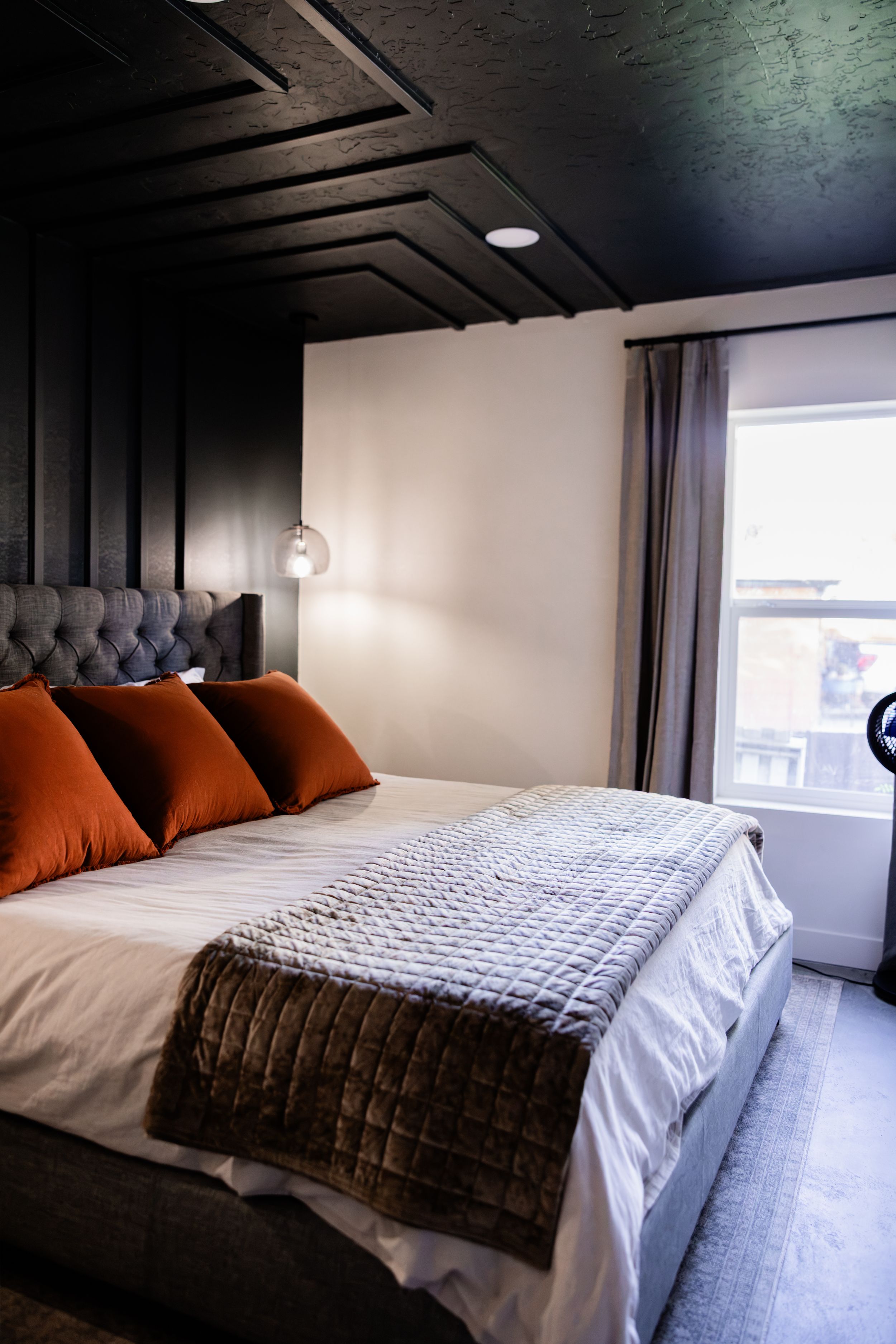 Elegant hotel bedroom with tufted headboard, burnt orange pillows, quilted throw, and dramatic dark ceiling with pendant lighting.