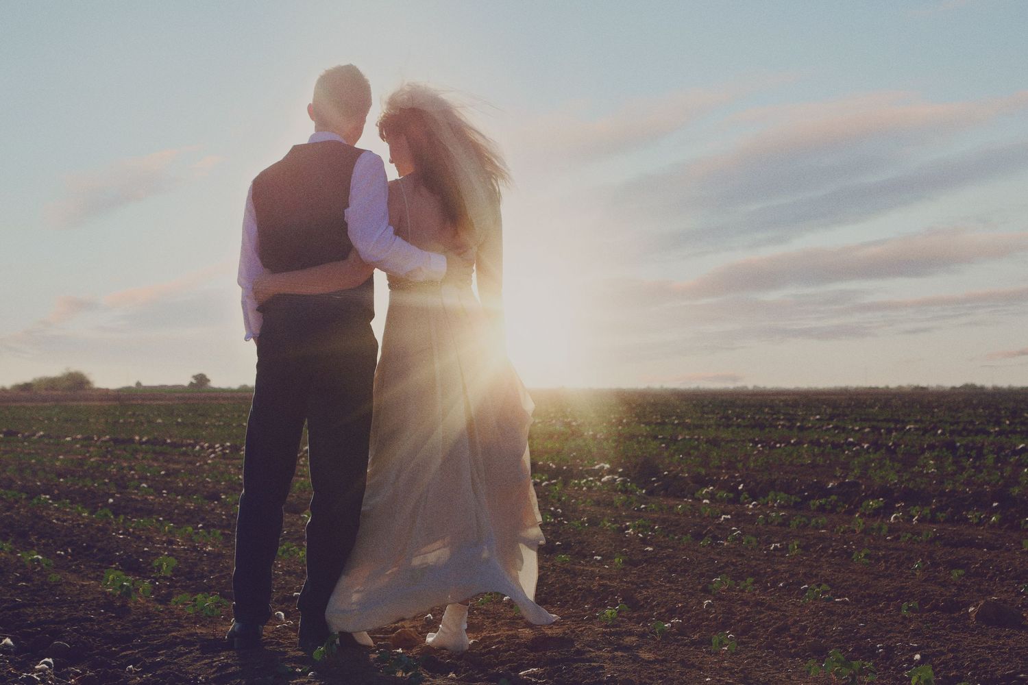 A couple in wedding attire embraces while facing a glowing sunset over an open field.