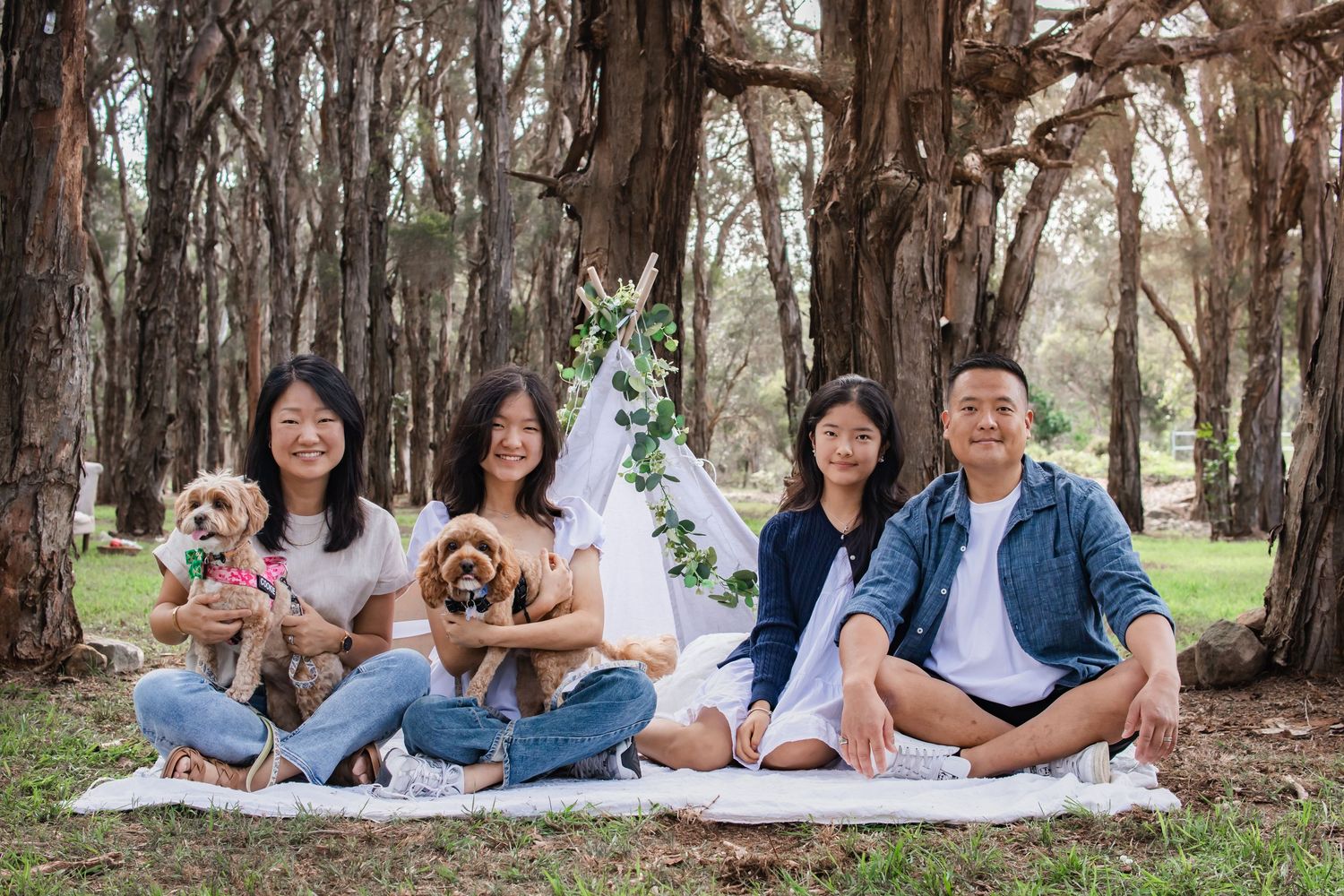 Family of four sitting on a blanket with two small dogs beside a white teepee tent and leaf garland in a sunny forest in Beaumont Hills.